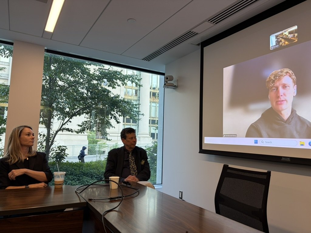Two people seated at a seminar-room table beside a window, with Winston Weinberg of Harvey filling the wall screen on a video call.
