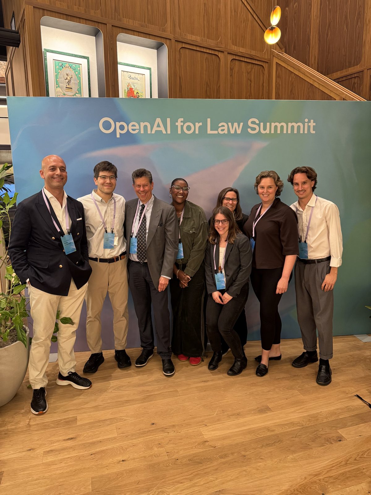 Eight people, lanyards on, in front of a backdrop reading &ldquo;OpenAI for Law Summit&rdquo;.
