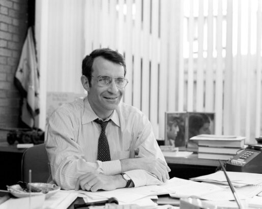 A black and white image of a man wearing a collared shirt and tie sitting behind a desk and sheaves of papers. He has glasses and smiles.