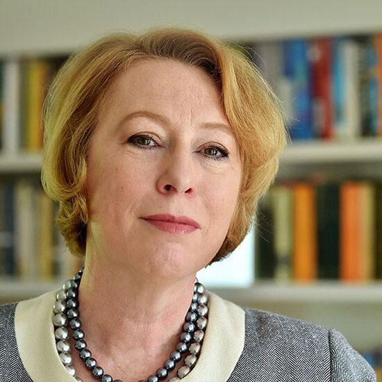 A white woman with short blonde hair and chunky necklaces peers at the camera in front of a bookcase.