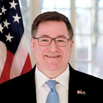 Man in glasses with a suit and blue tie in front of an American flag.