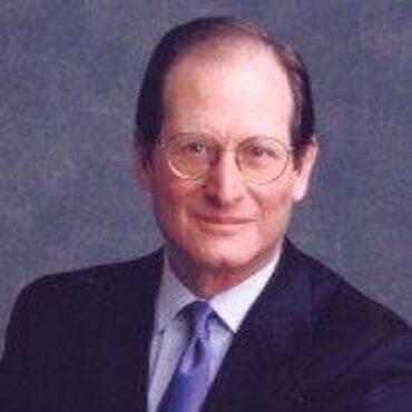 An older white man with a receding hairline and glasses looks out at the camera with a slight smile. He wears a suit and stands in front of a vague, blue background.
