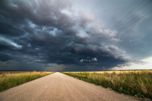 Dark storm clouds descend on a dirt road leading out into the distance. On the right, sun peaks through.