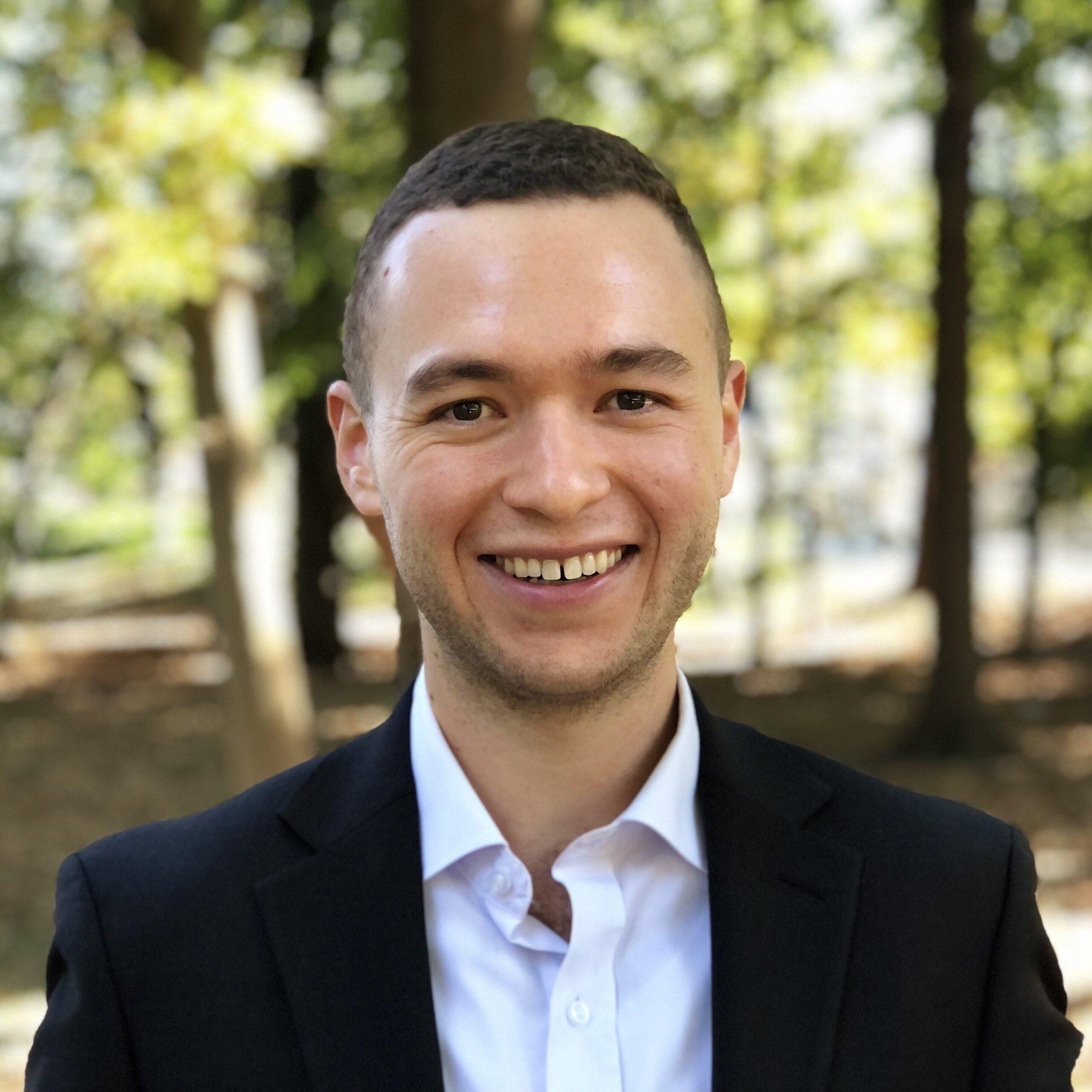 A young man with close cropped hair smiles in front of a forest; he wears a suit jacket and collared shirt.