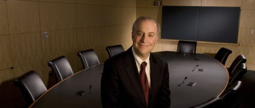 An older white man wearing a suit and tie stands in front of a conference table and leather chairs. His hands are clasped and he smiles faintly.