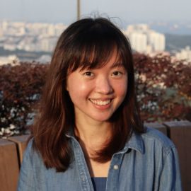 A young Asian woman with short brown hair and bangs smiles widely; behind her is a city scape. She wears a denim shirt.