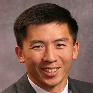 An Asian man wearing a suit smiles at the camera in front of a purple backdrop.