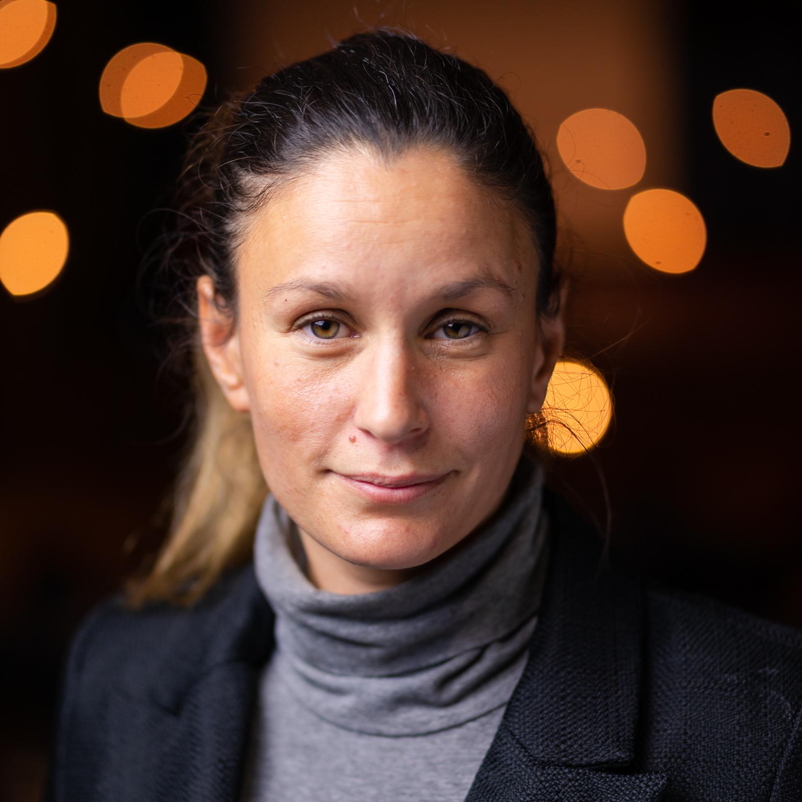 A white woman stares warmly out at the camera, wearing a turtle neck and a blazer. Behind her lights flash in the distance.