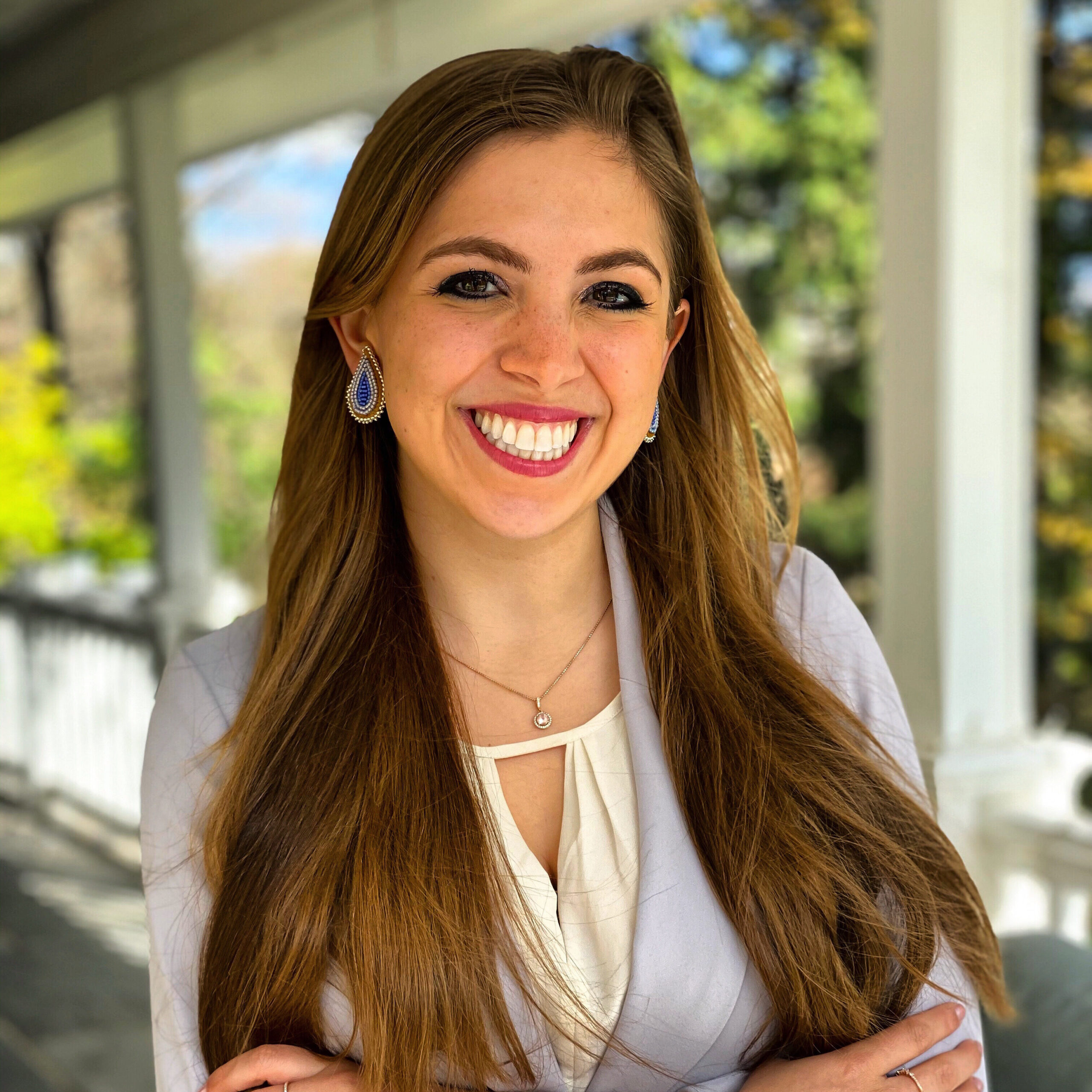 A young woman with very long brown hair smiles widely at the camera, wearing a light purple jacket and white blouse. She has big beaded earrings and red lipstick on and her arms are crossed.