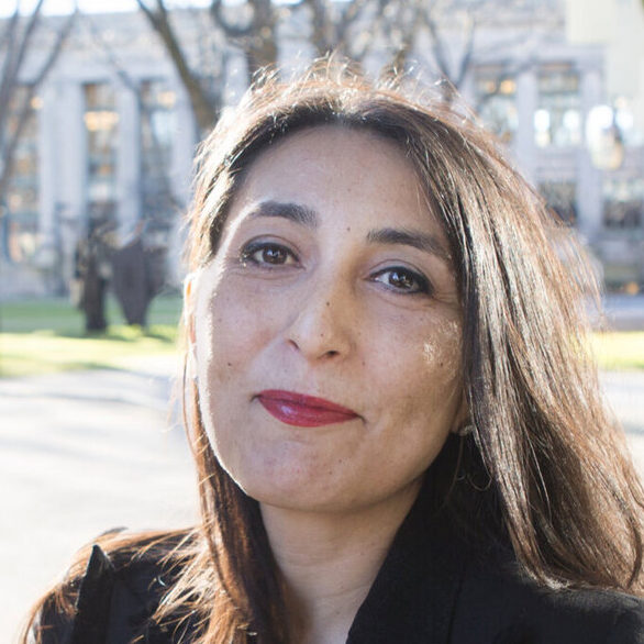 Woman wearing a wool peacoat looks into the sunlight. She wears red lipstick and has brown hair. She stands next to a glass wall and in front of a campus quad.