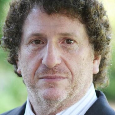 A white man with curly hair wearing a suit stares blankly at the camera.