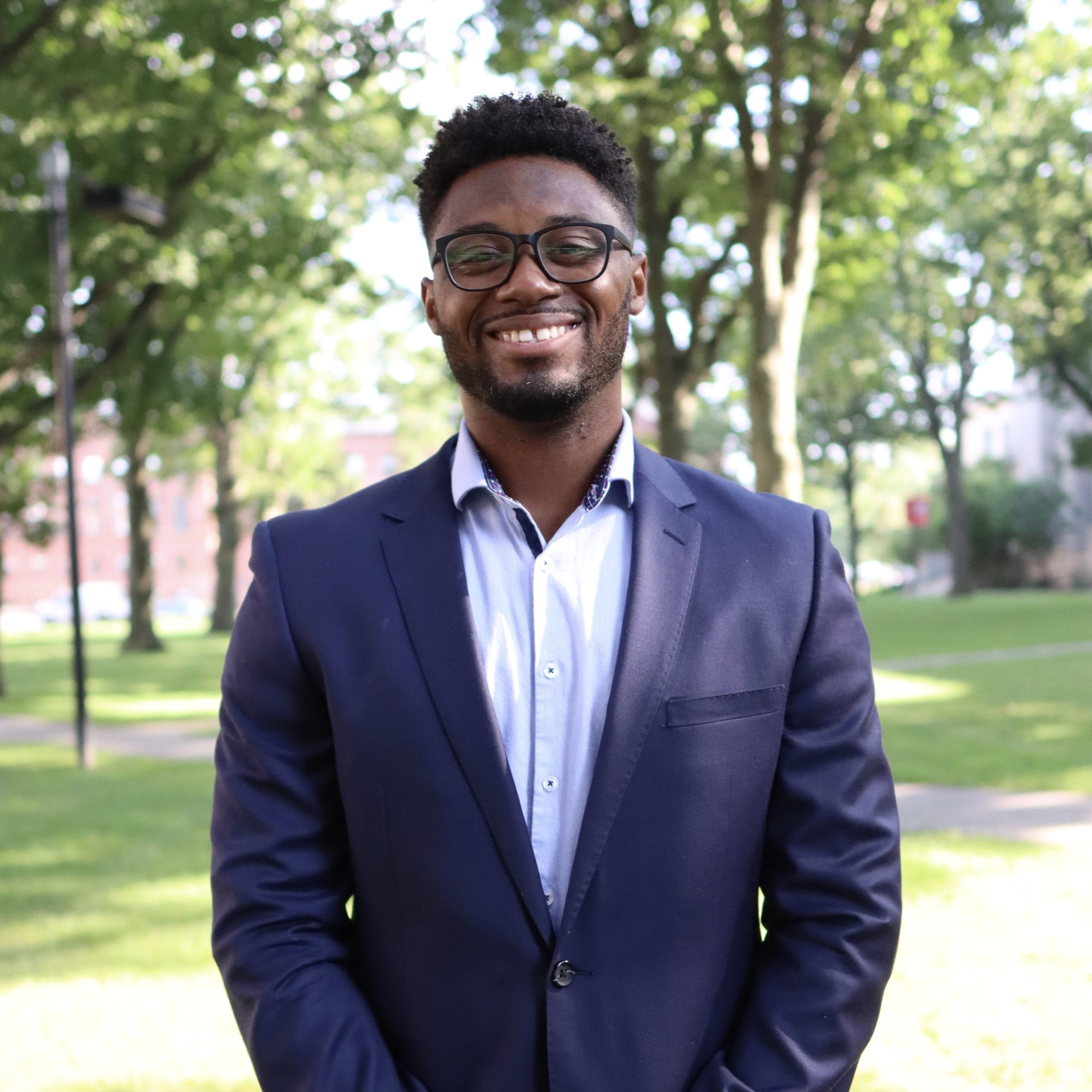 A young Black man wearing a blazer and button-down blue shirt smiles against the Harvard Yard backdrop. He has glasses and some facial hair. Trees dot the landscape behind him.