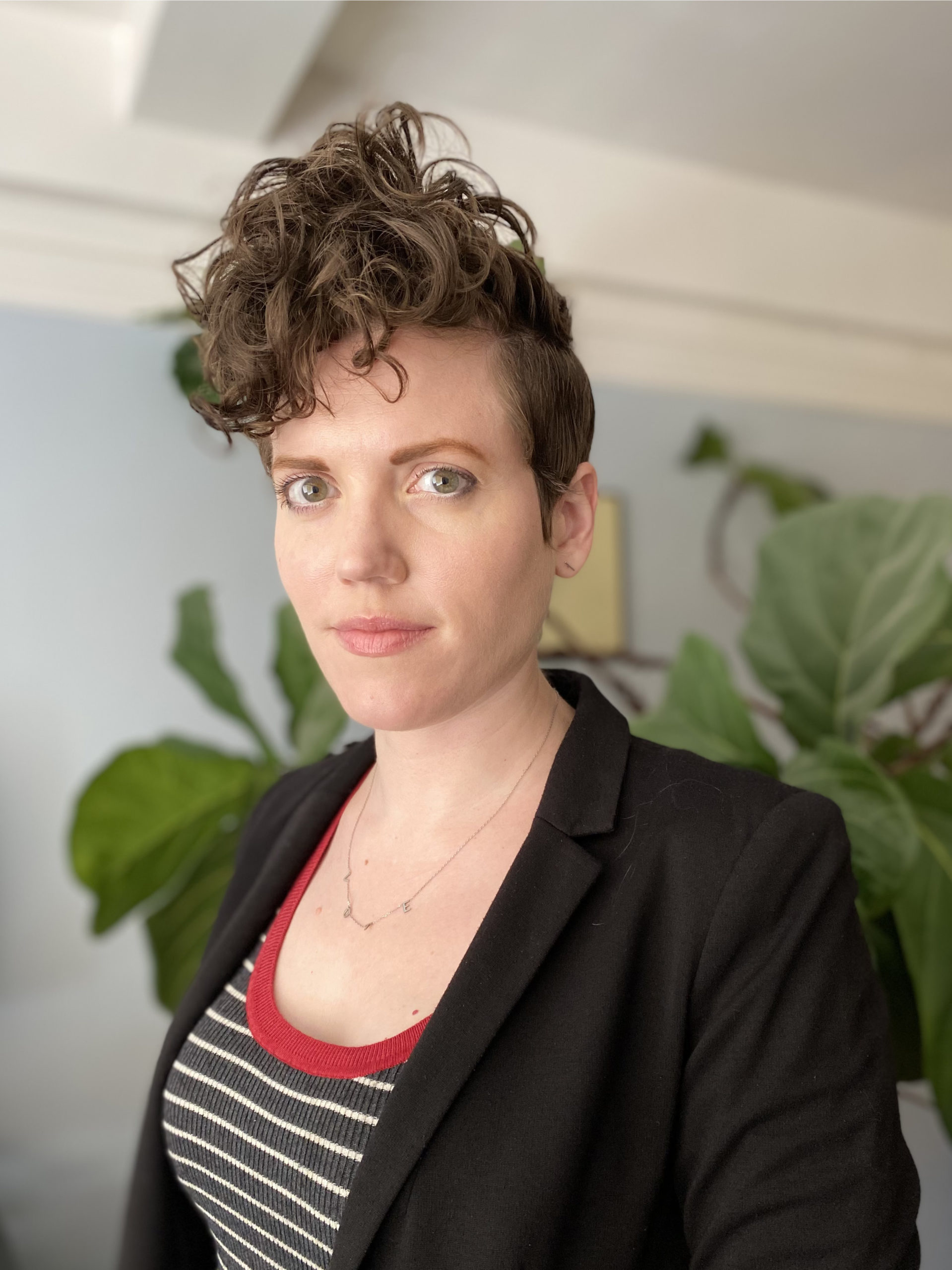 A white woman with short curly hair that falls above her forehead stares directly at the camera. She wears a black blazer over a striped top.