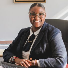 A young Black woman wearing a blazer and a tie sits behind an office desk and smiles.