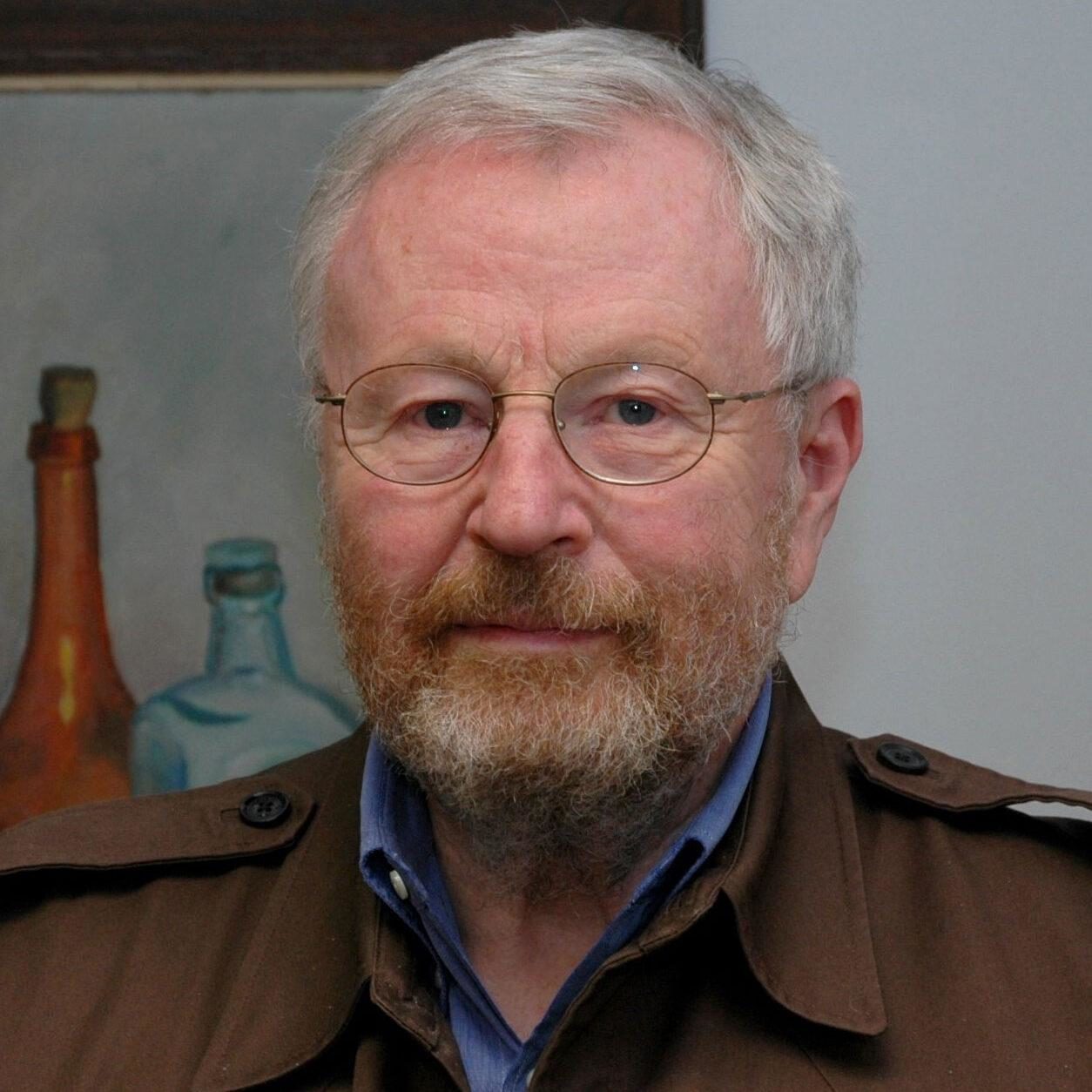 An older white man with a beard and glasses wearing a brown collered jacket stares out at the camera, in front of a still life of bottles.