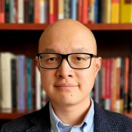 Asian man with glasses, mid-40s, sitting in front books.