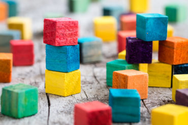 Colorful wooden blocks are scattered around a wood table. Some are piled on top of each other.