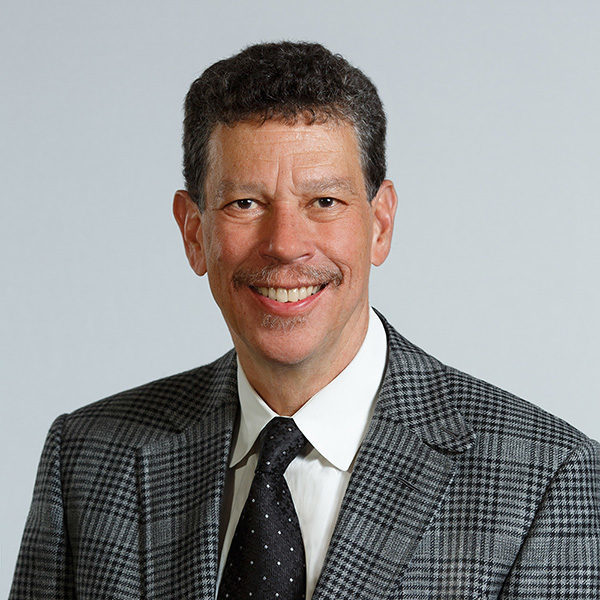 A man of about 65 with short curly hair wearing a checkered suit and a pocket square and tie smiles against a grey background