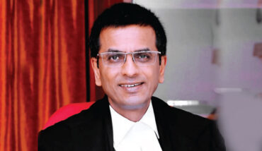 An Indian man wearing a judge's uniform and glasses sits in front of a red cloth.