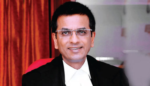 An Indian man wearing a judge's uniform and glasses sits in front of a red cloth.