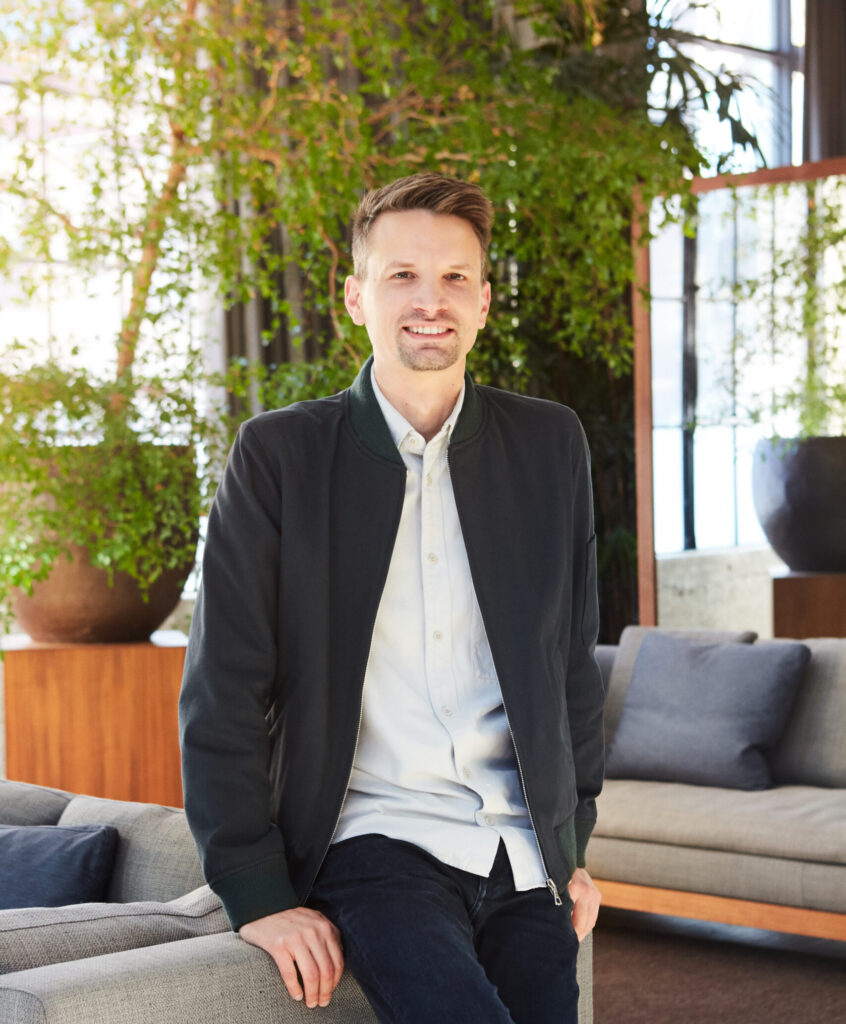 Image of Jason Boehmig leaning back against a couch in front of some greenery in an office building.