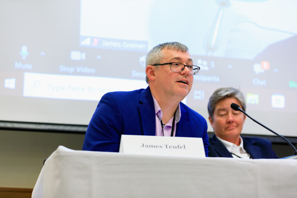 A man leans forward to talk into a microphone next to a woman watching on a panel table.