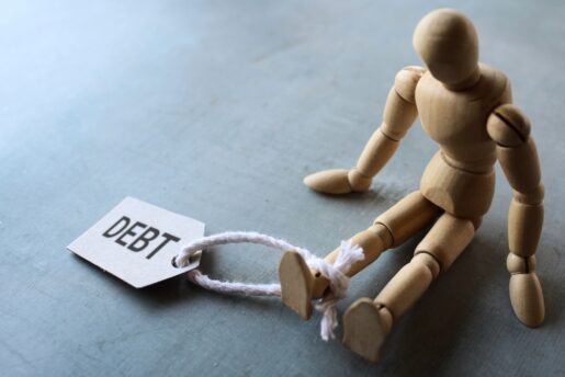 A wooden toy is slumped over with a tag around its ankle that says debt.
