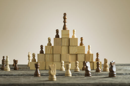 White and black chess pieces sit on a hierarchy of wooden blocks with one on top.